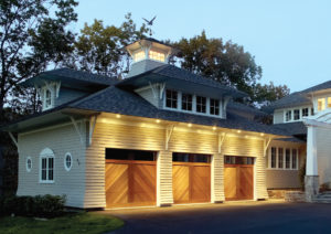 Three wood-grain garage doors with windows across top, on a large home at dusk, with under-eave lighting above the doors.